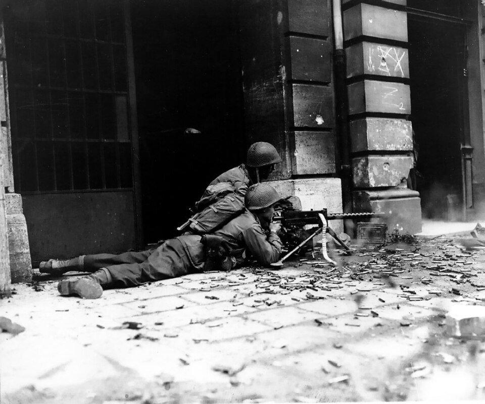 Two soldiers in steel helmets crouch and lie behind a machine gun in a stone doorway, spent cartridge cases scattered across the cobbles in front of them.