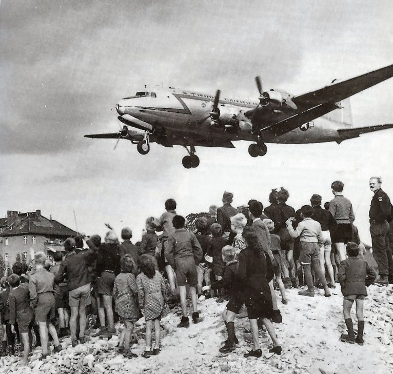 A four-engined transport aircraft coming in low over a crowd of children standing on a heap of rubble, several waving.