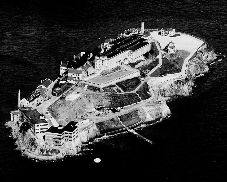 An aerial photograph of a rocky island in dark water, a long cell block and lighthouse along its spine and a fringe of docks and workshops at the near end.