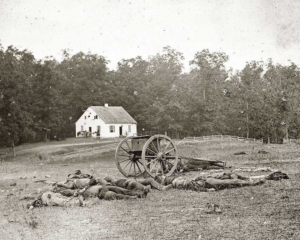 An early photograph of a bare field with an abandoned gun carriage and bodies laid in a row, a small white church among trees behind.