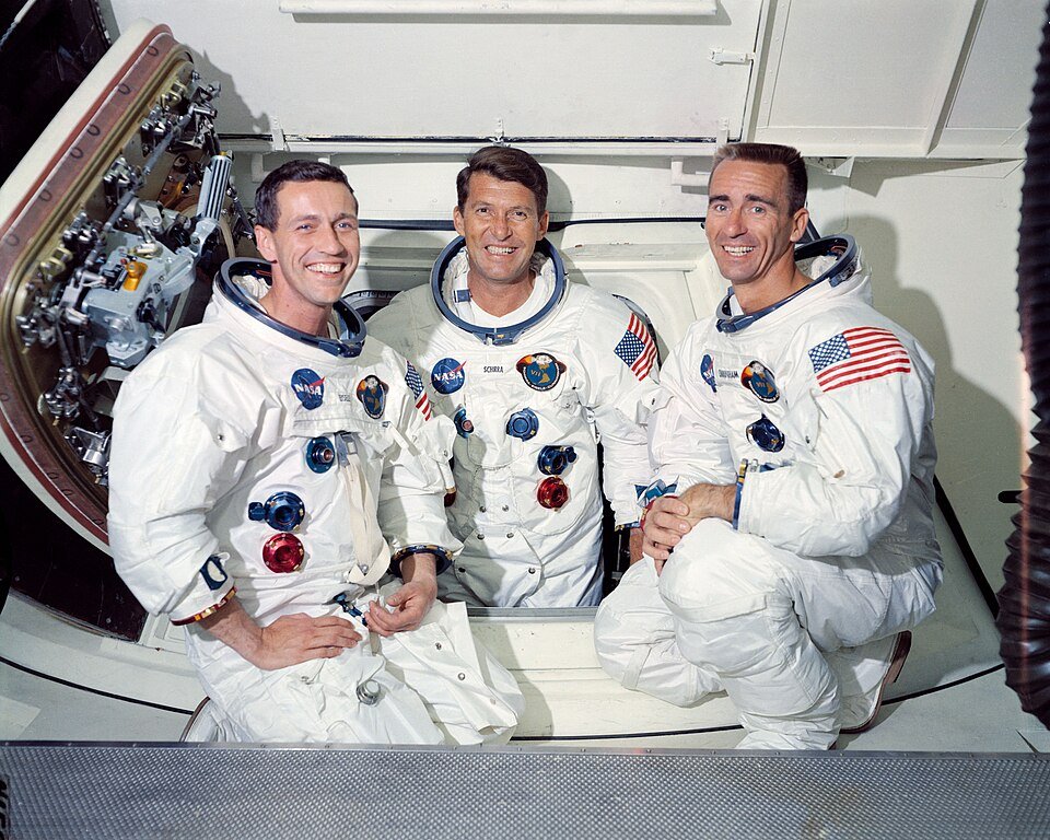 Three men in white spacesuits without helmets, sitting together in front of a spacecraft hatch, smiling at the camera.