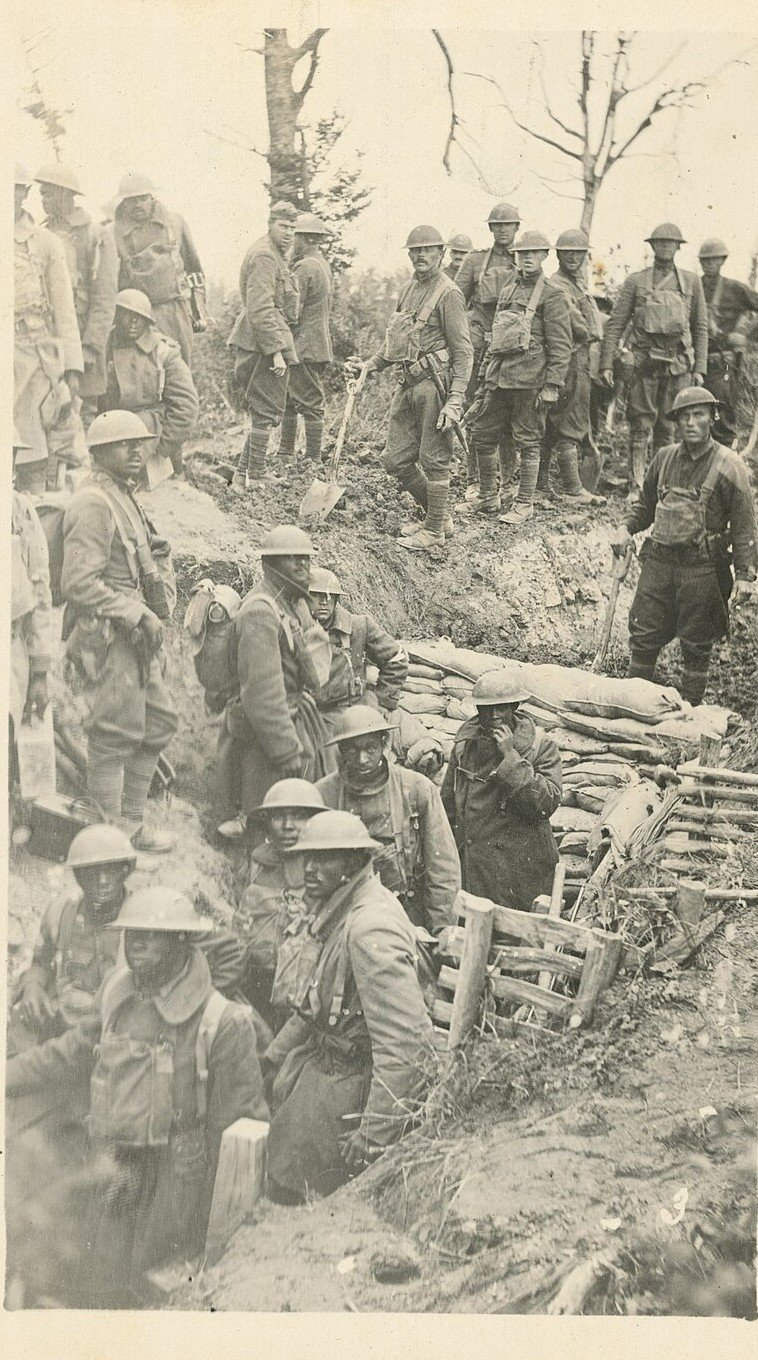 Soldiers in steel helmets crowded into a shallow trench among shattered trees, some standing on the parapet with shovels.