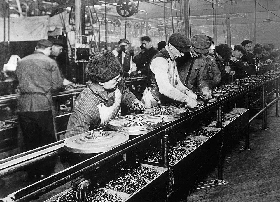 Men in flat caps and aprons working along a long moving rail, fitting parts to circular metal components, bins of small parts beside them and a crowded factory floor behind.