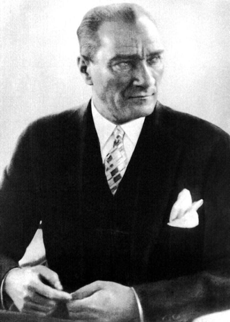 A black and white portrait of a man with pale eyes and swept-back hair in a dark three-piece suit and patterned tie, seated with his hands clasped.