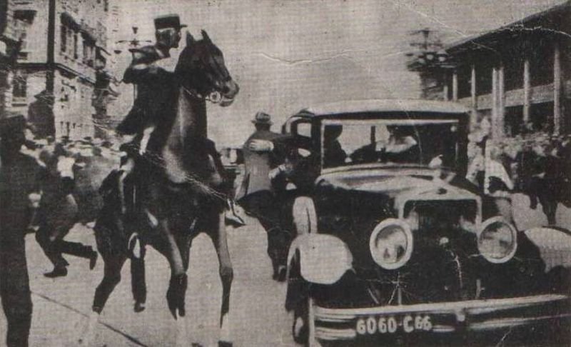 A grainy newspaper photograph of a mounted policeman rearing on his horse with a sabre raised, beside a large open car in a city street, with crowds scattering.