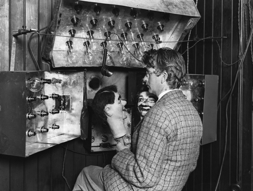 A man in a tweed jacket seen from behind, leaning into a home-made apparatus of metal panels studded with dozens of glass bulbs, holding a ventriloquist's dummy head in front of a lens.
