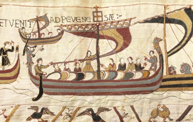An embroidered scene of a long open ship with a striped hull and a dragon prow, crowded with men and horses, with Latin lettering above.
