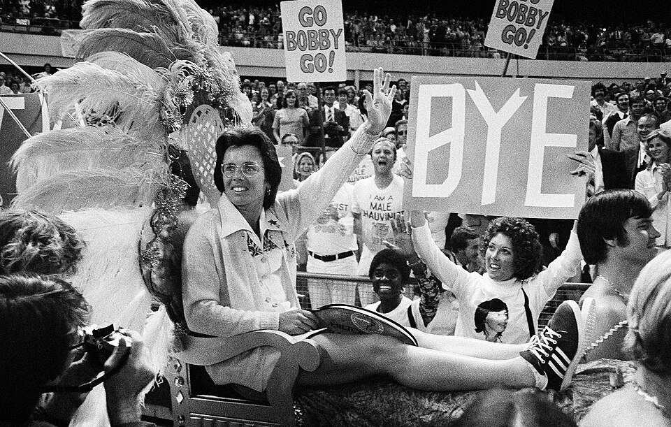 A woman in a tennis dress being carried into a packed arena on a decorated chair, waving, as spectators hold up handmade signs.
