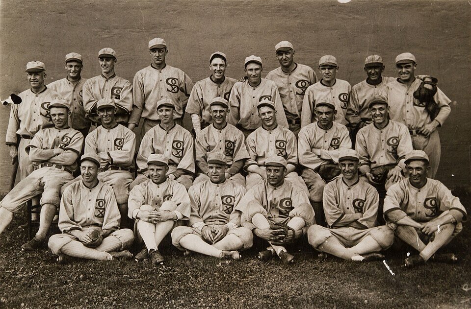 A sepia team photograph of about twenty baseball players in pale flannel uniforms with a scripted logo on the chest, arranged in three rows in front of a wall.