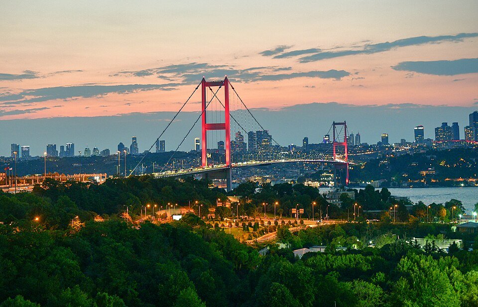 A long suspension bridge lit up at dusk spanning a wide strait, with a city skyline on the far shore and wooded hills in the foreground.