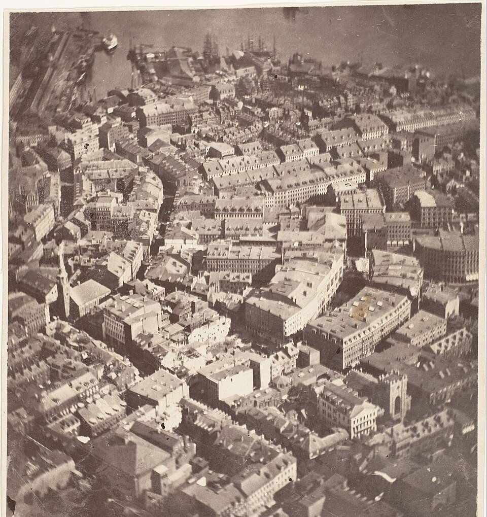 A sepia photograph looking steeply down on a dense grid of nineteenth century rooftops, streets and church spires, with harbour wharves and masted ships along the top edge.