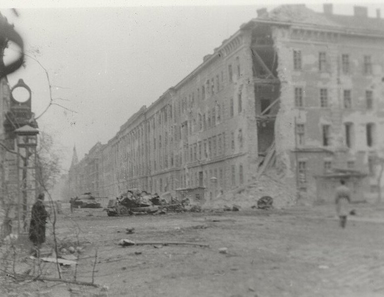 A wide city street with a long damaged apartment block down one side, wrecked vehicles standing in the road, debris across the cobbles and a few figures in the distance.