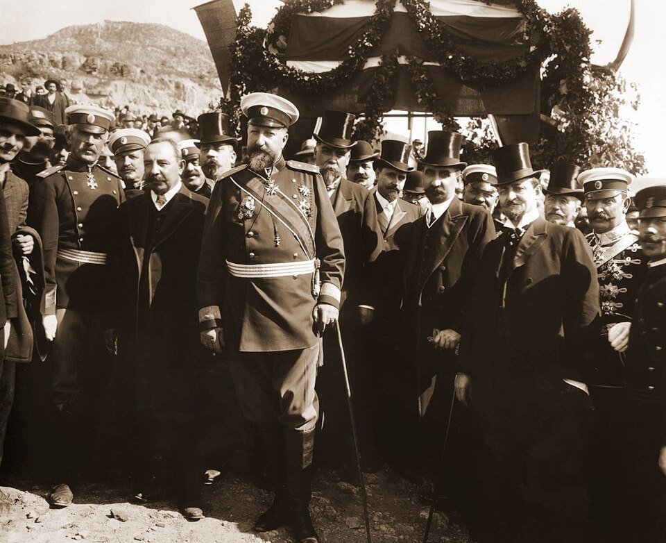 A sepia photograph of a stout bearded man in a military greatcoat and peaked cap standing in front of a garlanded platform, surrounded by officers and men in top hats.