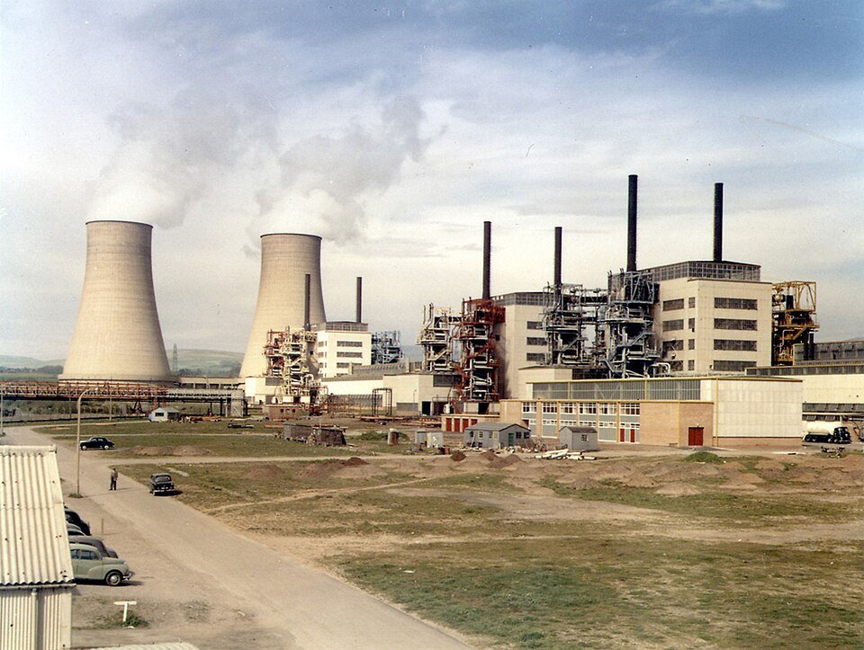 A colour photograph of an industrial site with two concrete cooling towers venting steam, tall chimneys and a concrete reactor hall, with 1950s cars parked on a track in the foreground.