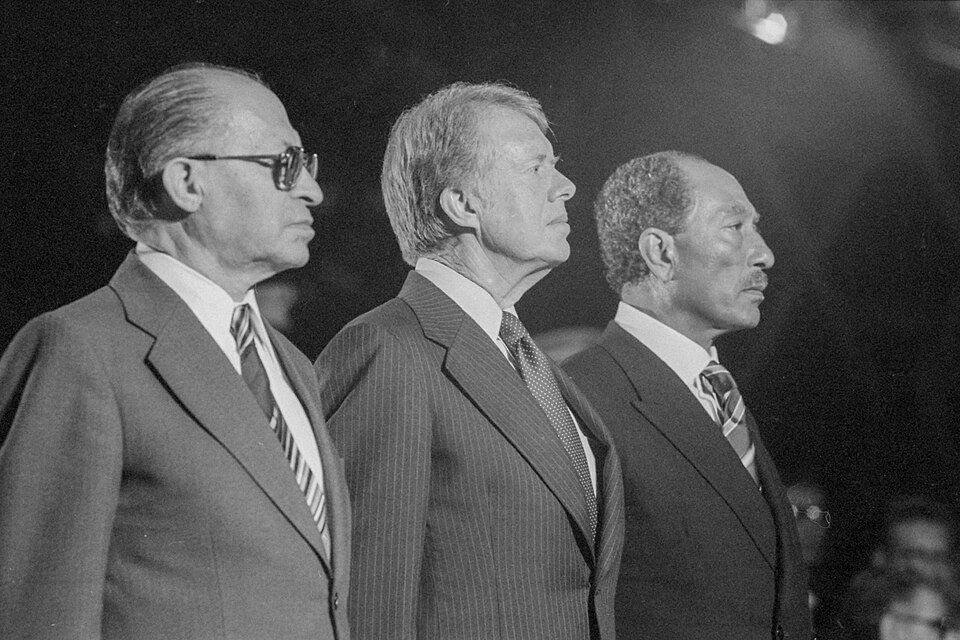 Three men in dark suits photographed in profile side by side, the middle one looking past the camera.