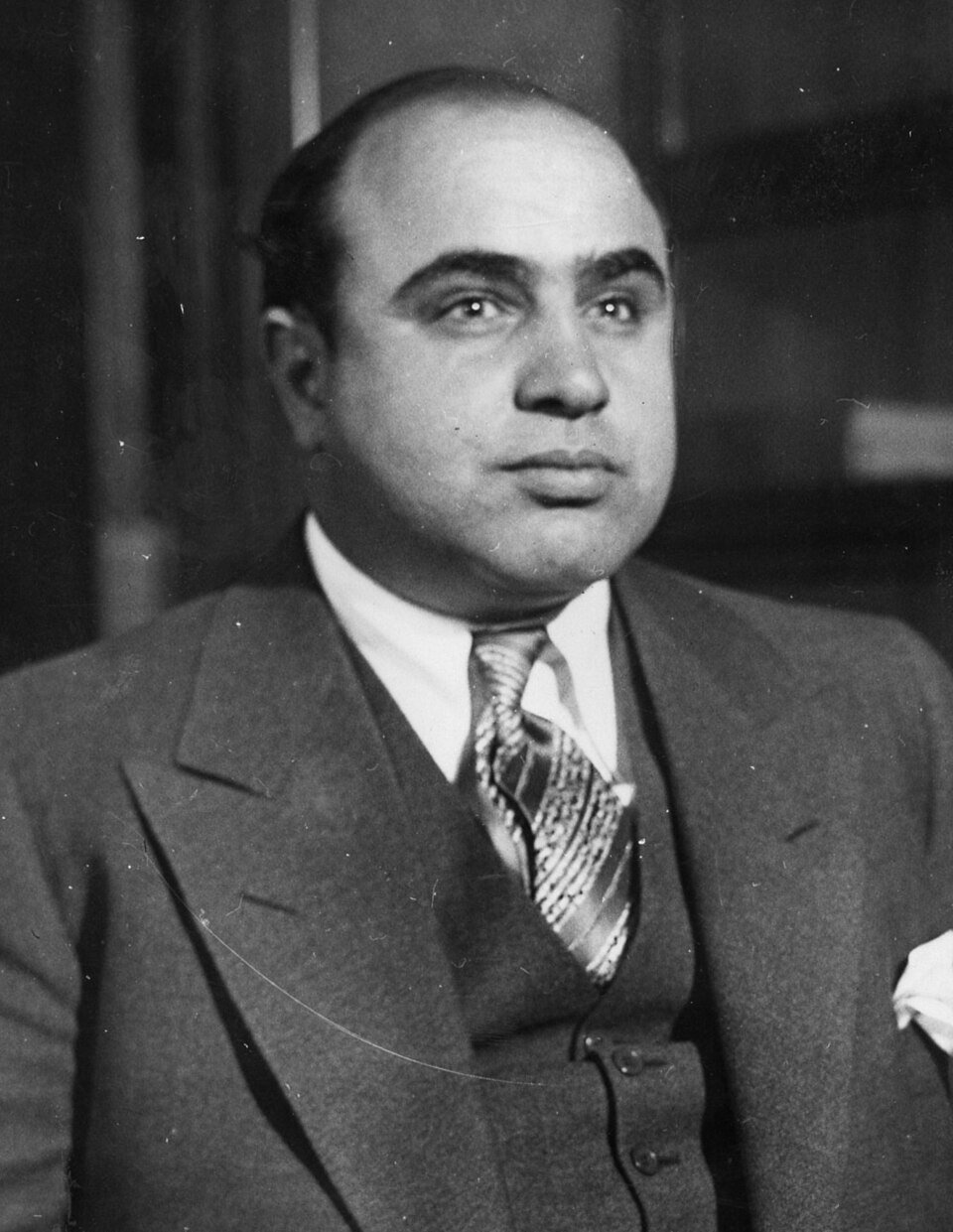 A black and white portrait of a heavy-set man in a three-piece suit and patterned tie, hair slicked back, looking steadily at the camera.