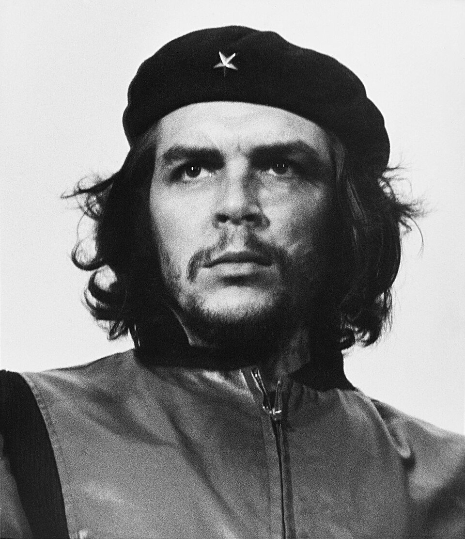A high contrast black and white portrait of a bearded man in a black beret with a star badge, long hair, looking upward past the camera.