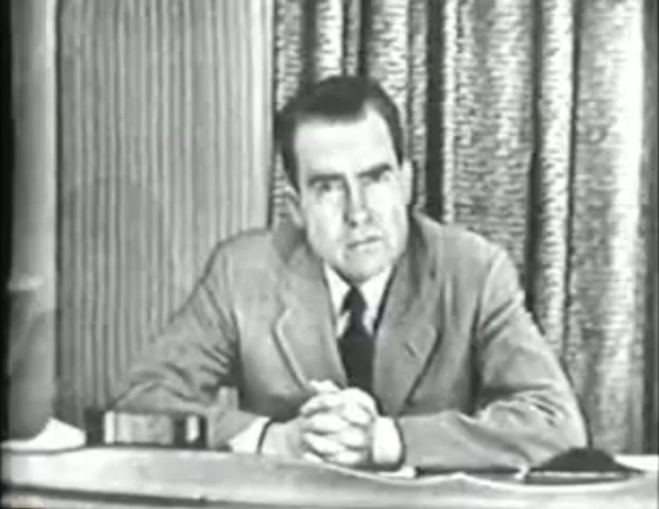 A grainy television still of a man in a suit sitting at a desk in front of heavy curtains, hands clasped, speaking to camera.