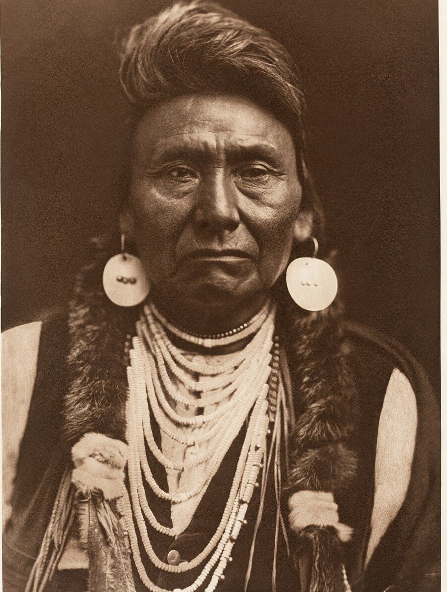 A sepia portrait of a man with swept-back hair wearing large disc earrings, many strands of shell beads and a fur-trimmed robe, looking directly at the camera.