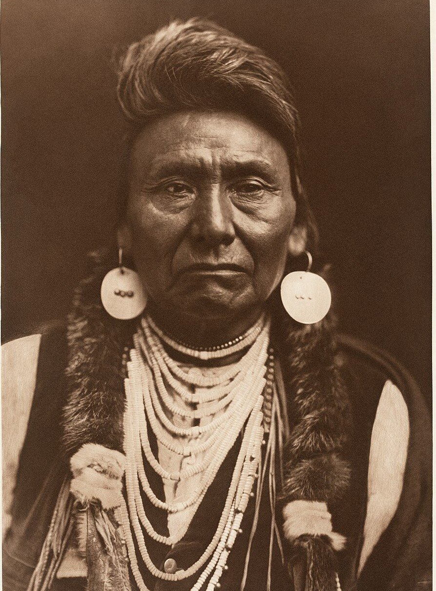 A sepia portrait of an older man with swept-back hair, large shell earrings and many strands of beads, wearing fur over his shoulders.