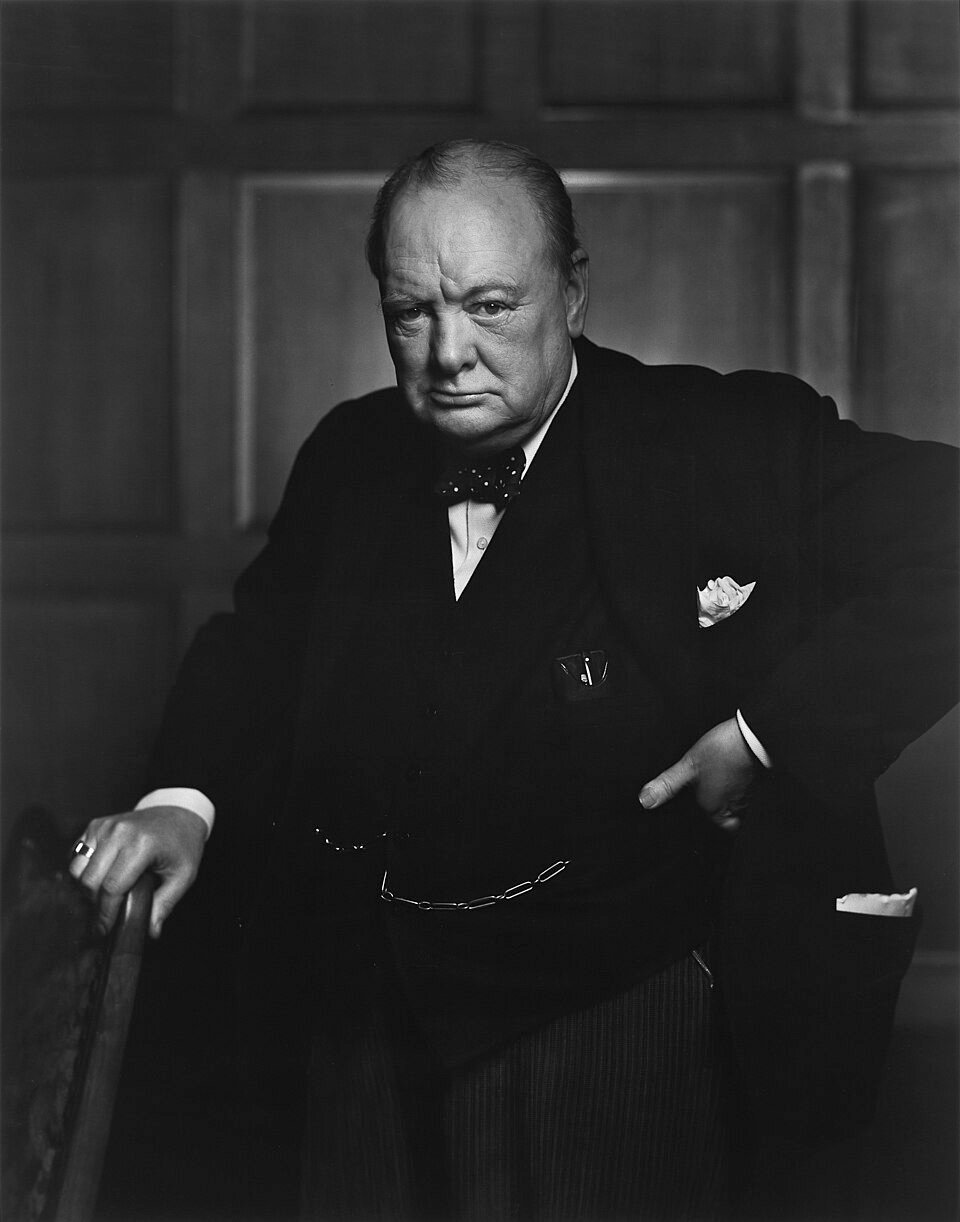 A black and white portrait of a heavy-set elderly man in a dark suit and bow tie, scowling directly at the camera with one hand on the back of a chair.