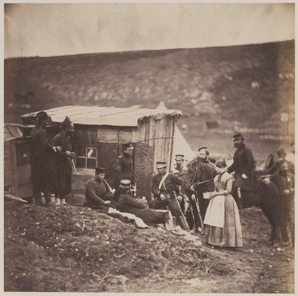 A sepia photograph of officers and soldiers gathered outside a rough timber hut on a bare hillside, a woman in an apron among them and a horse being held on the right.