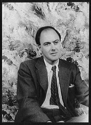 Black and white portrait of a dark haired man in a tweed jacket seated against a mottled backdrop.
