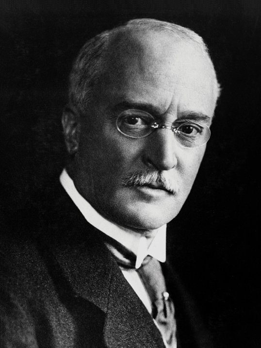 A formal photograph of a bald man with a heavy moustache and pince-nez in a dark suit.