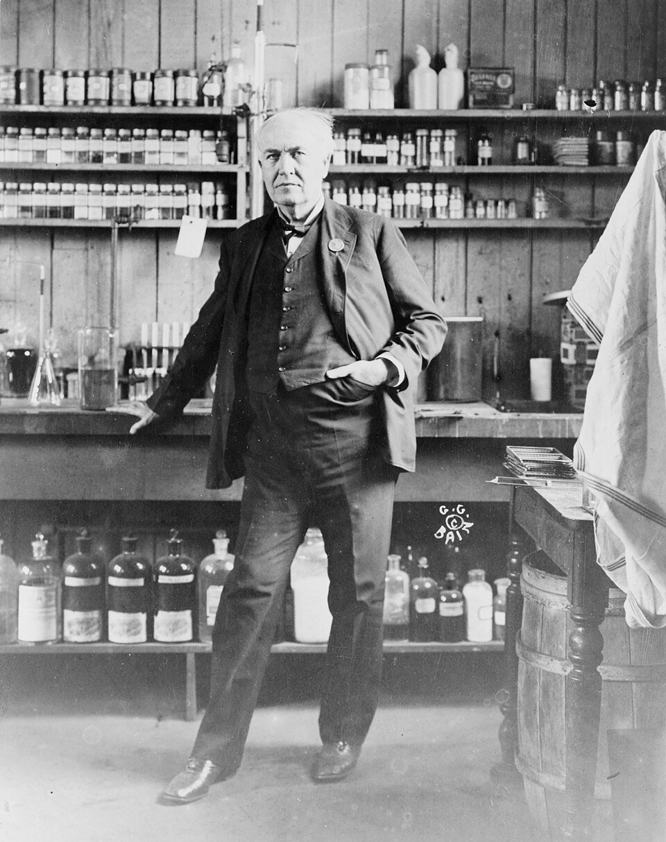 A photograph of an elderly man in a rumpled suit standing in a laboratory lined with shelves of glass bottles and chemical apparatus, hands in his pockets.