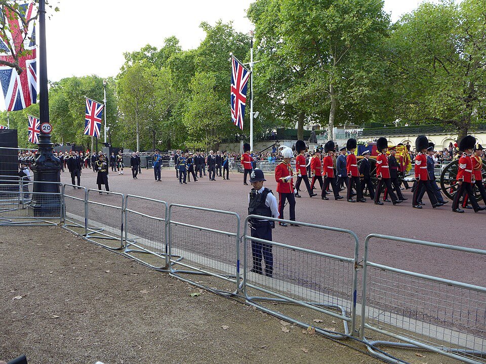 A state procession along a flag-lined avenue, with guardsmen in red tunics and bearskins escorting a gun carriage.