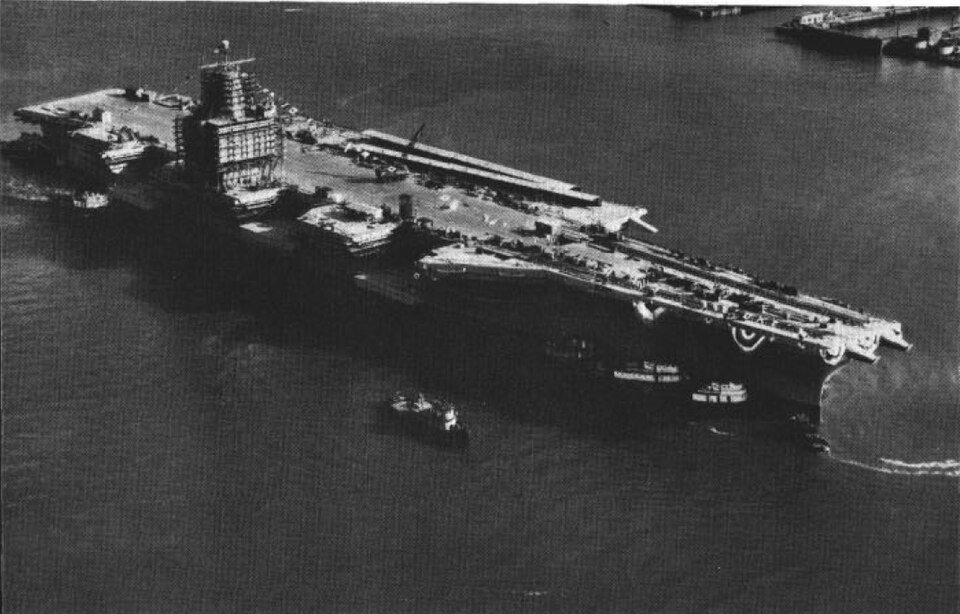 An enormous flat-decked warship being manoeuvred by tugs in a river shortly after launch.