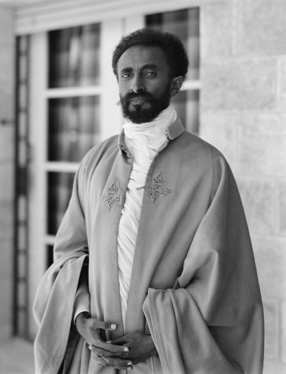 A formal photograph of a bearded man in an embroidered cape and white robe standing on a veranda, hands folded, looking past the camera.