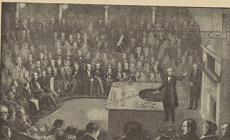 An engraving of a man demonstrating an experiment at a bench before a packed tiered lecture theatre.