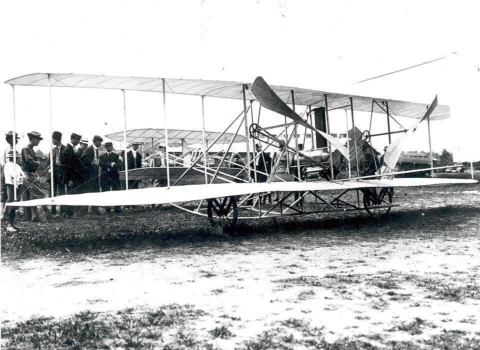 An early biplane of wood, wire and fabric standing on a grass field with a line of men in hats watching from behind it.