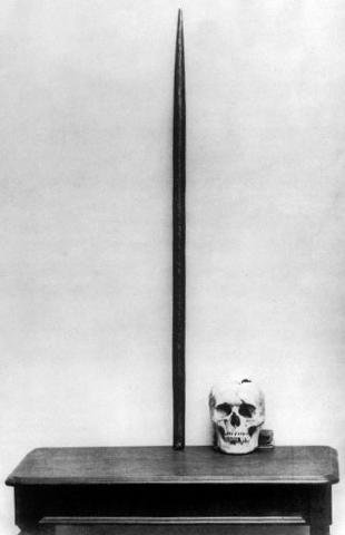 Black and white photograph of a human skull beside a long pointed iron rod standing upright on a table.
