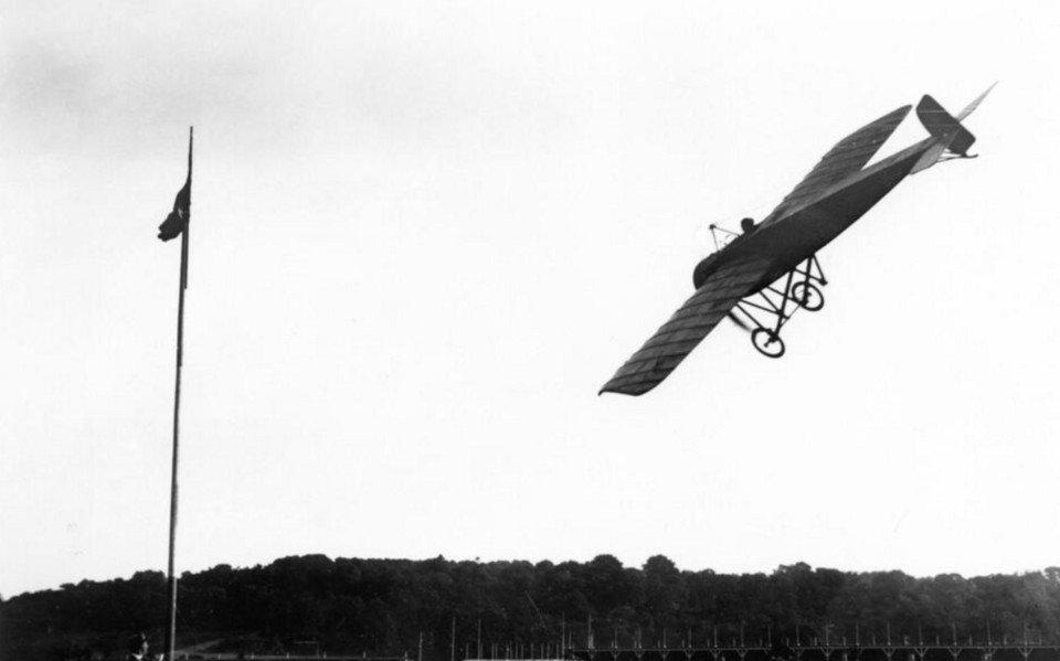 An early monoplane of wood and fabric banking steeply against a pale sky above a line of trees.