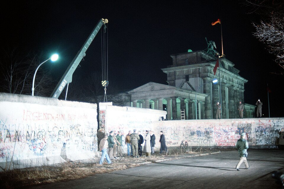 A night scene of a graffiti-covered concrete wall with a crane lifting one section clear, a floodlit classical gateway topped by a chariot behind it, and guards standing on the wall.
