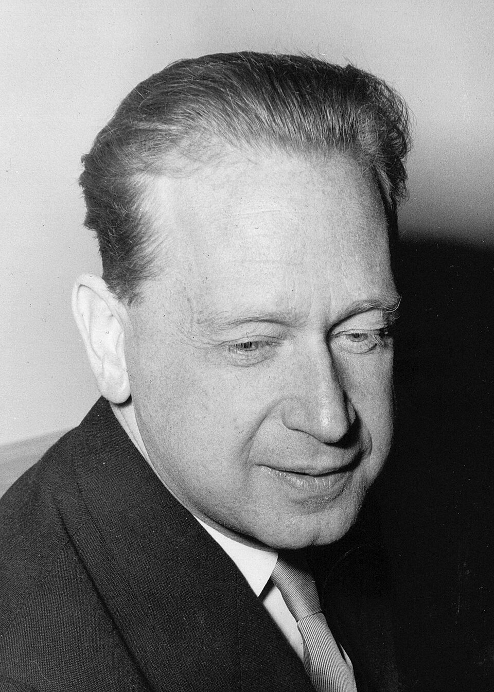 A black and white portrait of a man in a dark suit looking downwards with a slight smile.