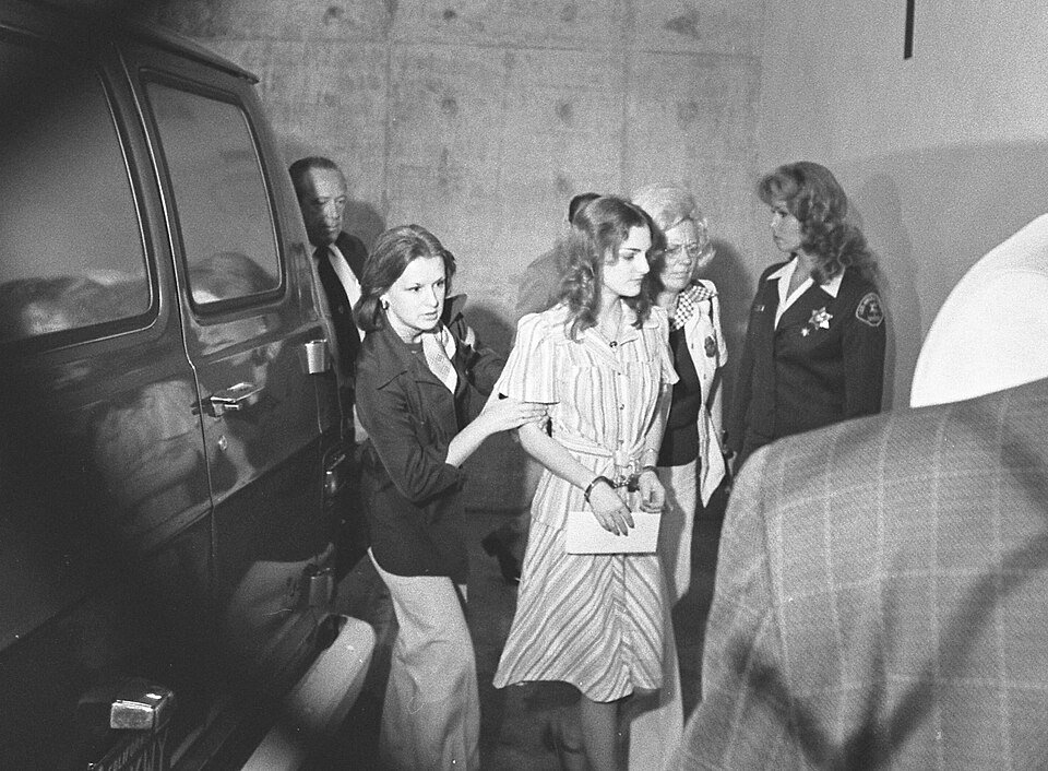 A young woman in a striped dress and handcuffs being led towards a van by two women in uniform jackets.