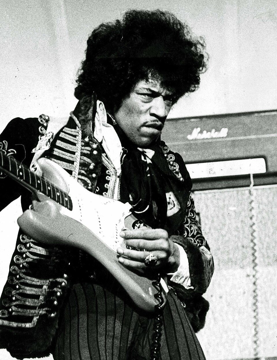 A black and white photograph of a guitarist with a large afro in a braided military jacket, holding a white electric guitar in front of an amplifier stack.