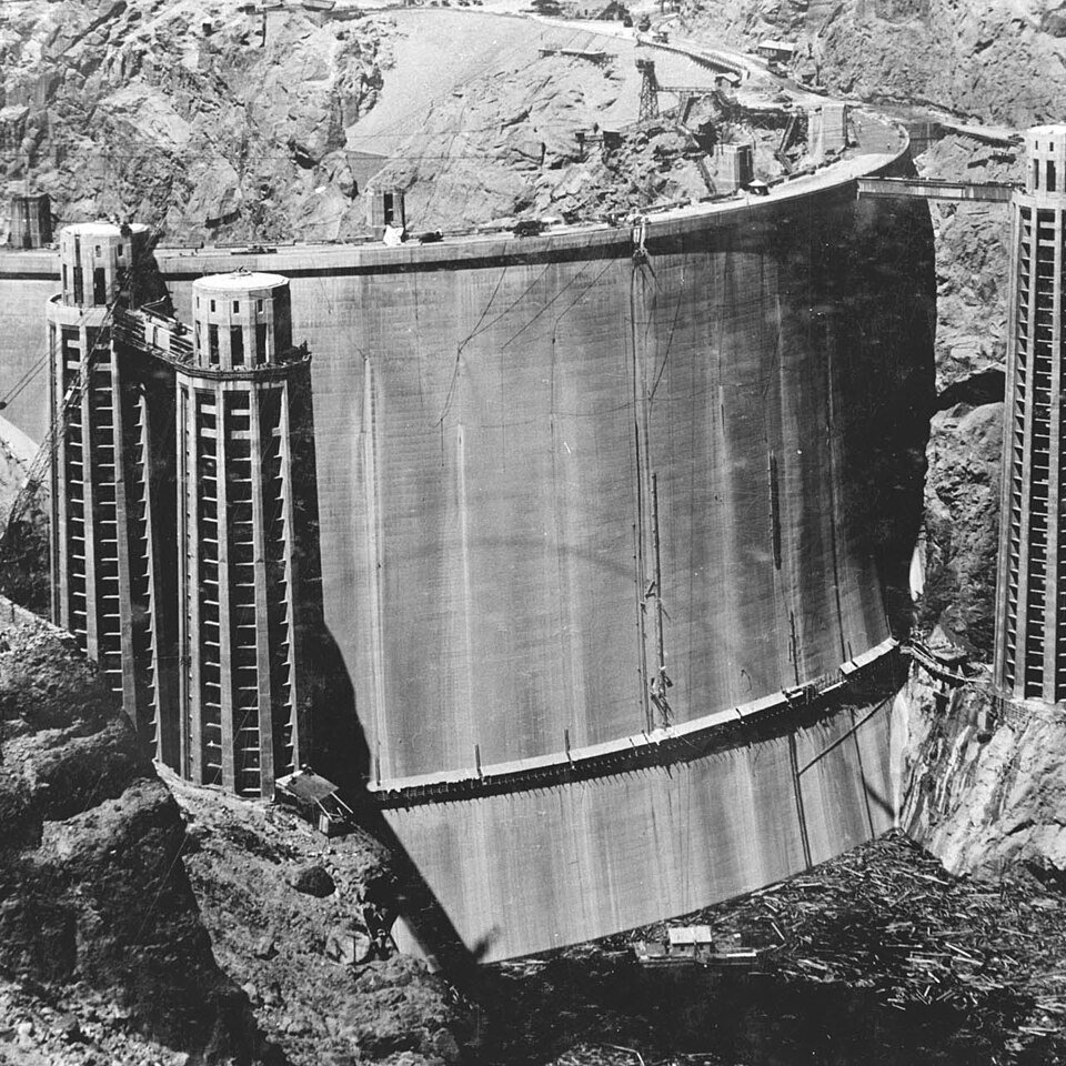 A vast curved concrete dam wall wedged between rock canyon walls, with four cylindrical towers along its crest.