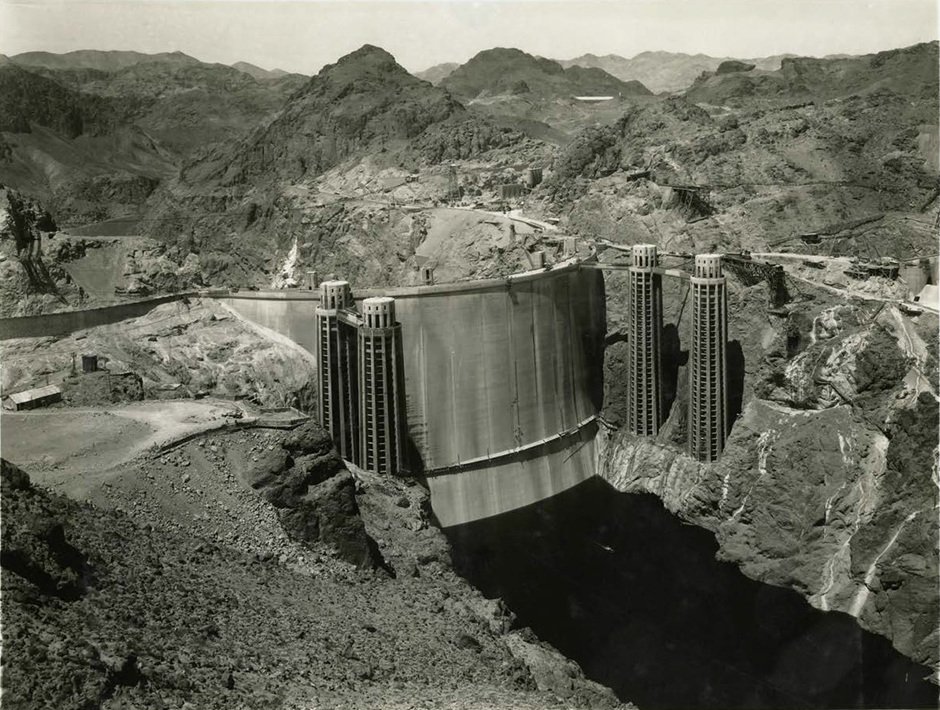 A period photograph of a huge curved concrete dam wedged between bare rock canyon walls, four tall intake towers along its crest and construction roads cut into the hillsides.