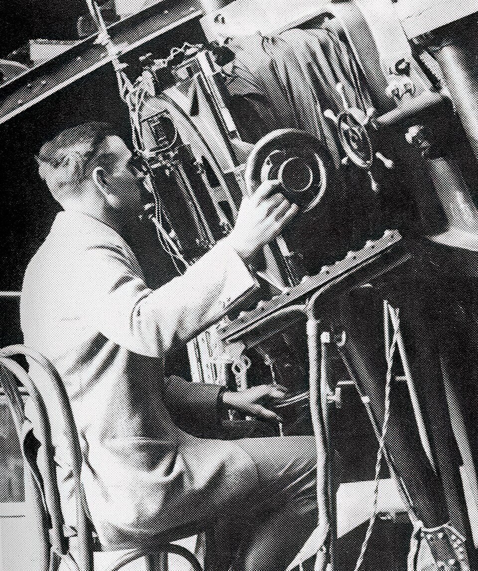 A man in shirtsleeves seated at the eyepiece of a huge telescope among girders and cables.