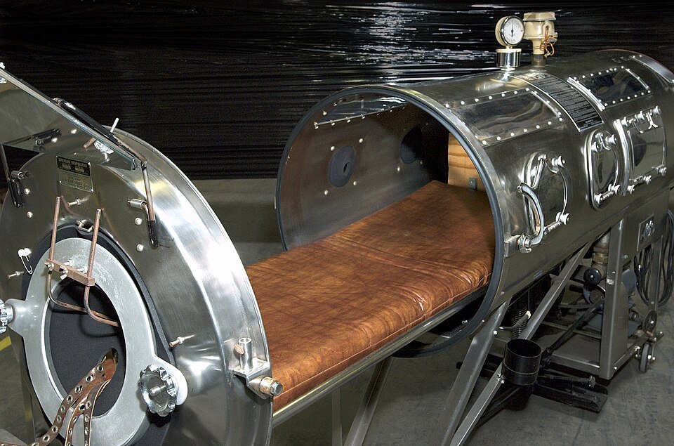 A polished metal cylinder on a wheeled frame, open at one end with a sliding bed and a padded collar, gauges and valves along the top.