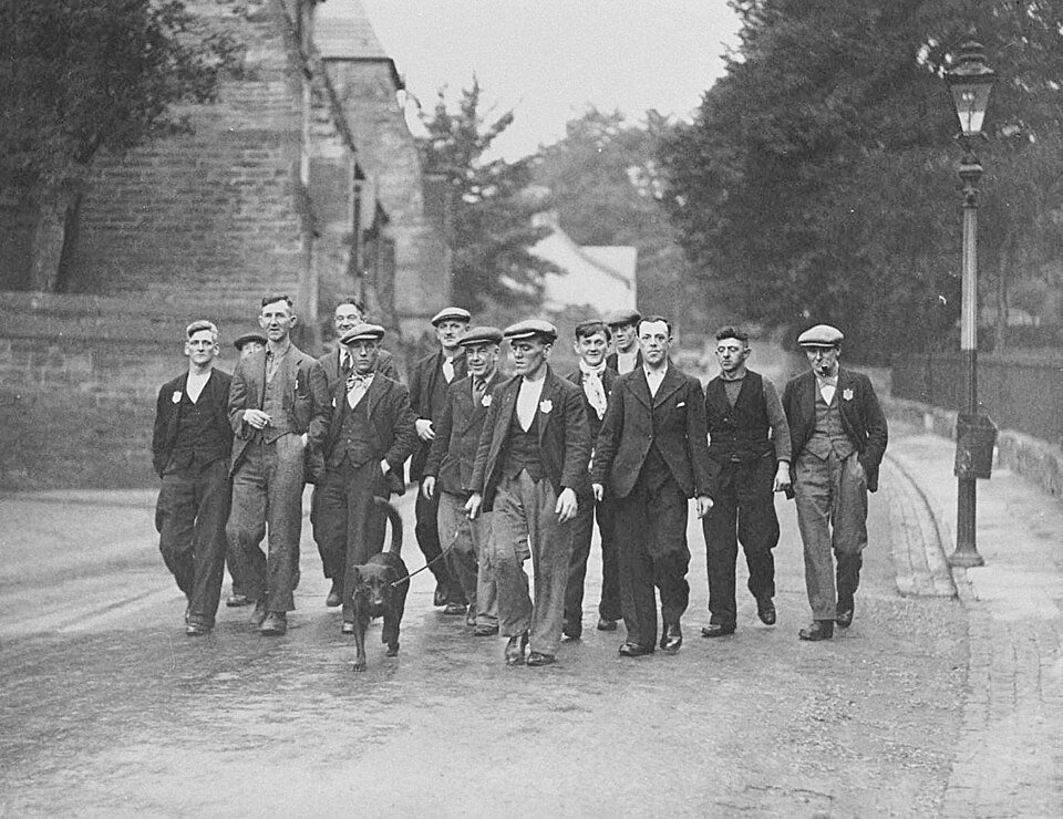 A group of men in flat caps, waistcoats and jackets walking down a road towards the camera, one holding a small dog on a lead, a stone wall and gas lamp behind them.