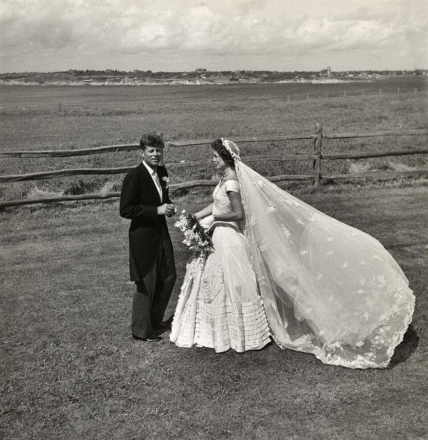 Black and white photograph of a bride with a long lace veil and a groom in morning dress standing in a field.