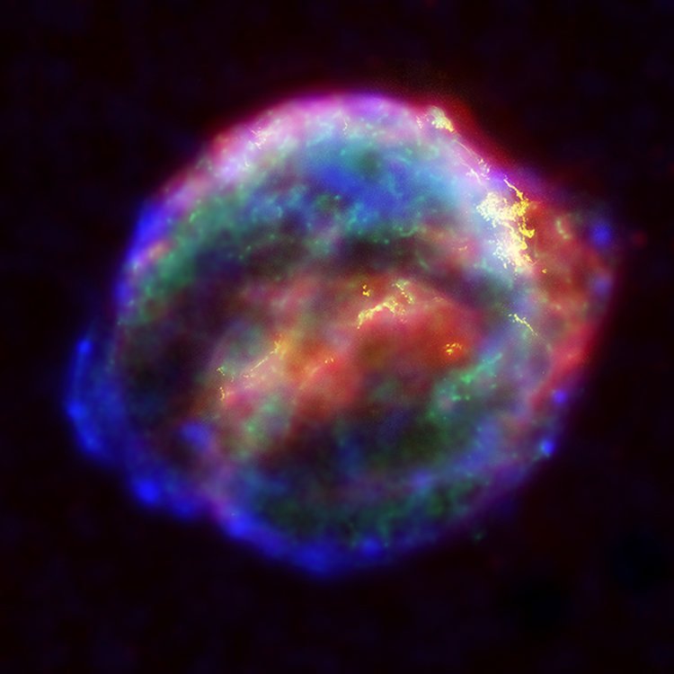 A false-colour astronomical image of an expanding shell of gas in pink, blue, green and gold against a dark field.