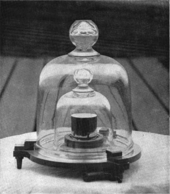 A small dark metal cylinder standing under two nested glass bell jars on a polished base.