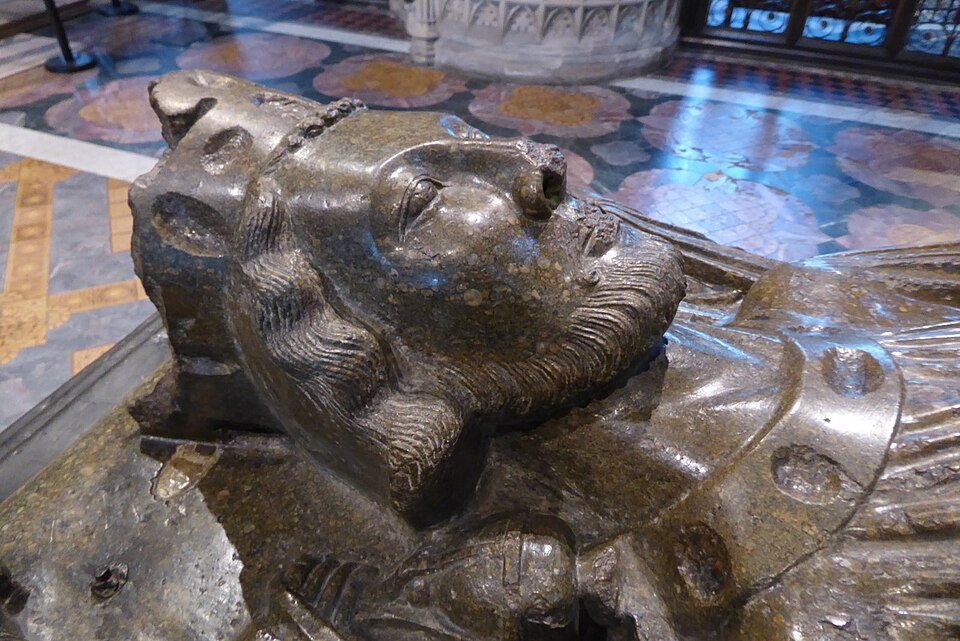 A close-up of a medieval tomb effigy carved in dark mottled stone, showing a crowned and bearded king lying in state, with a patterned floor beyond.