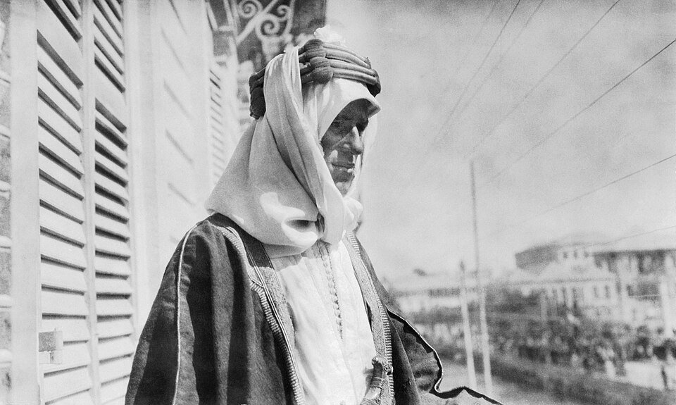 A man in Arab robes and headdress standing on a balcony with shutters behind him, a large crowd filling the street below.